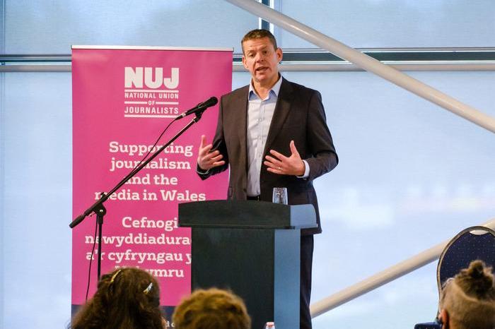 Rhun ap Iorwerth wearing a suit standing behind a lectern with a pink NUJ banner to his left.