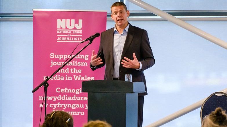Rhun ap Iorwerth wearing a suit standing behind a lectern with a pink NUJ banner to his left.