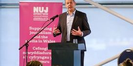 Rhun ap Iorwerth wearing a suit standing behind a lectern with a pink NUJ banner to his left.