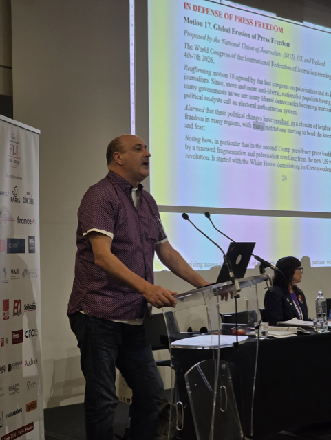 Gerry Curran, wearing a blue shirt, speaks behind a lectern with text on a screen in the background.