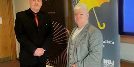 Gerry Curran wearing a black suit stands next to Gerard Cunningham wearing jeans and a grey blazer with an NUJ banner in the background.