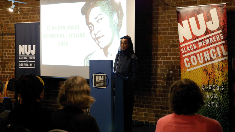 Nesrine Malik stands behind a blue podium with a projected image of Claudia Jones to her left and an NUJ Black Members' Council banner to her right.