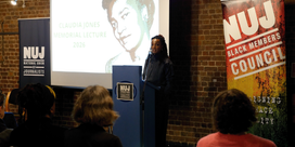 Nesrine Malik stands behind a blue podium with a projected image of Claudia Jones to her left and an NUJ Black Members' Council banner to her right.