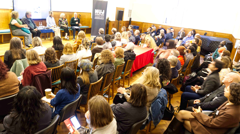 A dimly lit room full of seated people with a panel of speakers, a TV screen, and a green NUJ vertical banner.