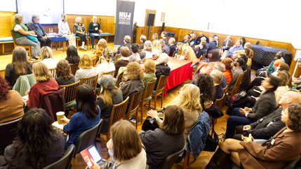 A dimly lit room full of seated people with a panel of speakers, a TV screen, and a green NUJ vertical banner.