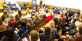 A dimly lit room full of seated people with a panel of speakers, a TV screen, and a green NUJ vertical banner.