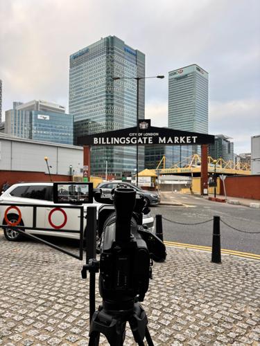 A camera pointed in the direction of a gate with the words 'Billingsgate Market' on it and grey skyscrapers in the background.