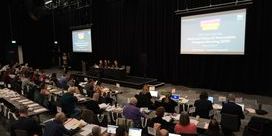 A dark conference room with delegates seated at chairs and desks with two large projected screens against black curtains.
